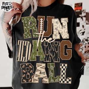 May include: A black t-shirt featuring the text RUN THE DANG BALL in large, block-style letters. Each letter is filled with various football-themed patterns, including helmets, footballs, field markings, and checkered designs in green, brown, and white.