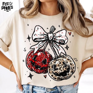 Puede incluir: Camiseta color crema con un dise&ntilde;o caprichoso de dos cerezas con estampado de leopardo y un lazo con detalles de cereza. El dise&ntilde;o incluye estrellas y corazones. El logotipo de la marca "FES PHIES" est&aacute; en la esquina superior izquierda.
