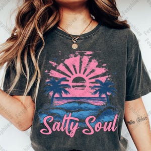 May include: A dark grey t-shirt with a pink and blue sunset graphic and palm trees. The text "Salty Soul" is printed in pink below the graphic.