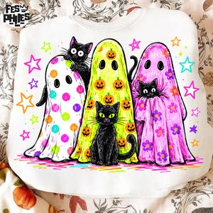 May include: A white sweatshirt featuring a graphic print of three ghosts and two black cats. One ghost has colorful polka dots, one has orange pumpkins, and one has purple flowers. The design includes colorful stars and the text FES PHIES.