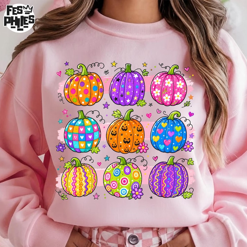 Op de afbeelding: Een lichtroze sweatshirt met een grafische print van negen pompoenen in diverse kleuren en patronen, waaronder stippen, sterren, bloemen, hartjes en strepen, gerangschikt in een raster met kleine sterretjes en hartjes eromheen.