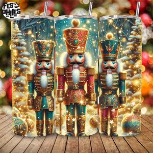 May include: Three stainless steel tumblers with a Christmas nutcracker design. The tumblers feature a festive design with a winter scene and a detailed nutcracker soldier in red, teal, and gold. The tumblers are decorated with gold accents and a straw.