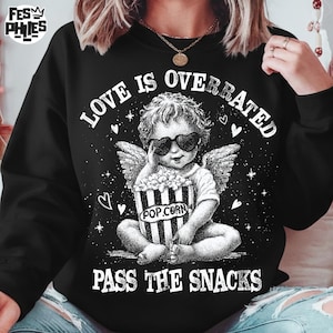 May include: Black sweatshirt with a graphic of a cherub wearing heart-shaped sunglasses, holding a popcorn bucket. Text reads "LOVE IS OVERRATED PASS THE SNACKS". The design includes wings, stars, and hearts.