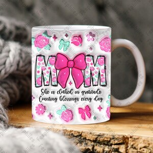 May include: White ceramic mug with a pink bow and the word "MOM" in pink letters. The mug has a floral pattern with pink roses and green leaves. The text "She is clothed in gratitude Counting blessings every day" is printed below the bow.