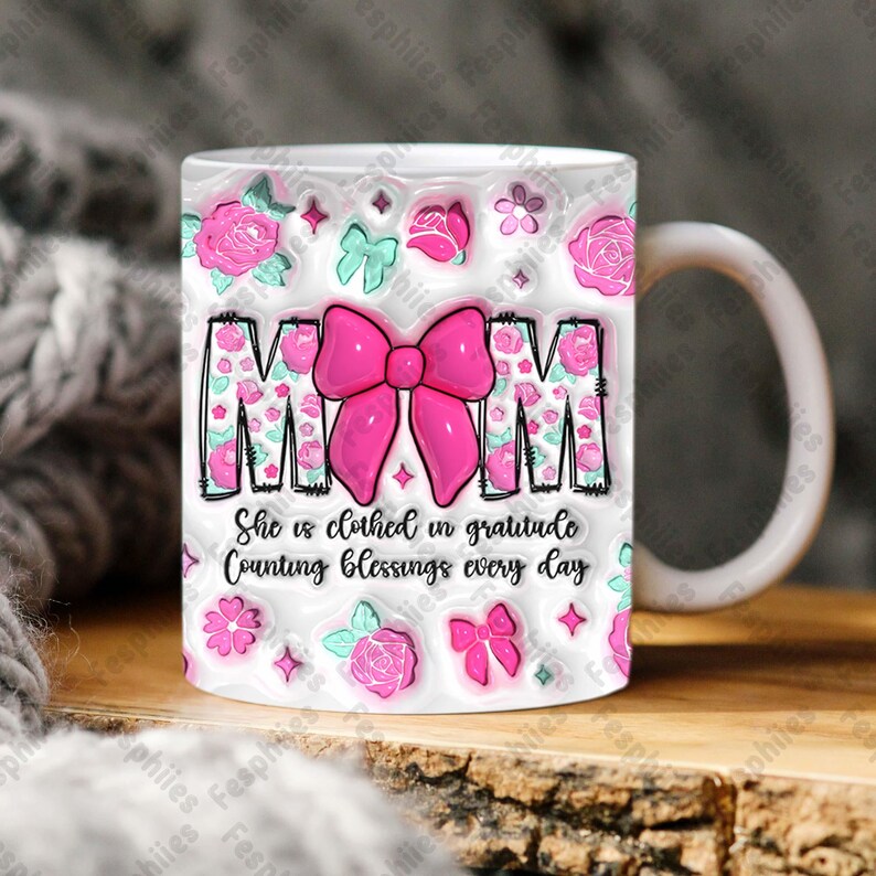 May include: White ceramic mug with a pink bow and the word "MOM" in pink letters. The mug has a floral pattern with pink roses and green leaves. The text "She is clothed in gratitude Counting blessings every day" is printed below the bow.