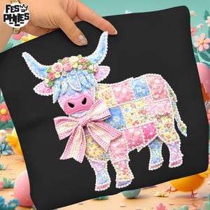 以下が含まれることがあります： 黒い正方形のクッション。パッチワークの牛のデザイン。牛は青い角、ピンクの鼻、花の冠があります。大きなピンクと白のギンガムチェックのリボンが首を飾っています。クッションは人が持っています。