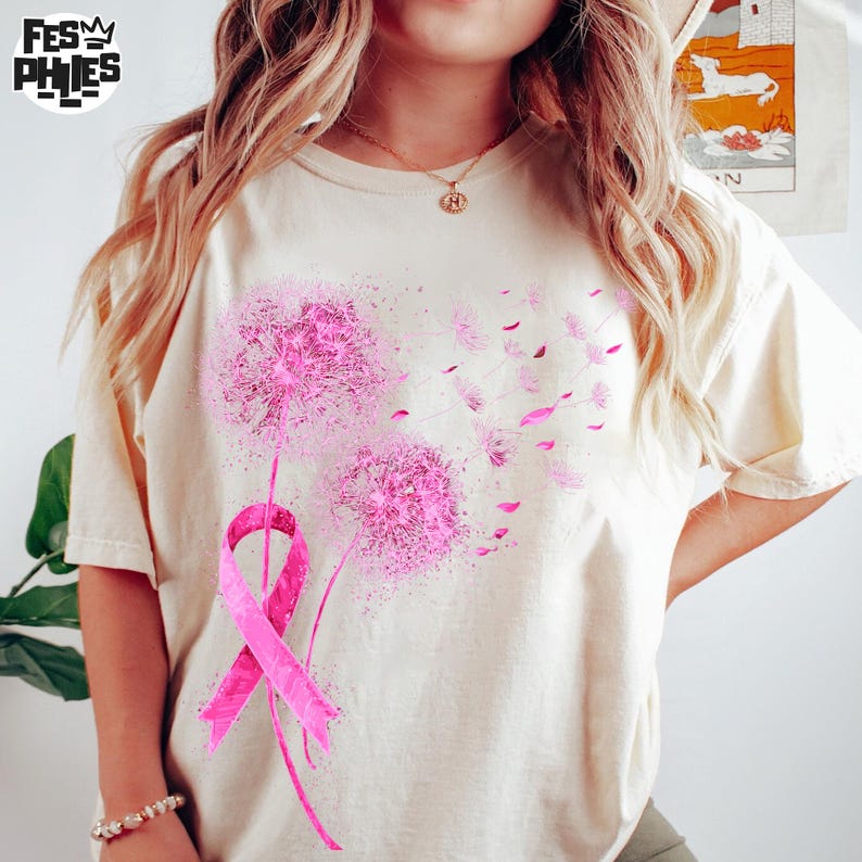 May include: Cream-colored t-shirt featuring a pink dandelion design with a pink ribbon. The dandelion's seeds are blowing away. The shirt has short sleeves and a crew neck. The logo "FESY PHIES" is in the upper left corner.