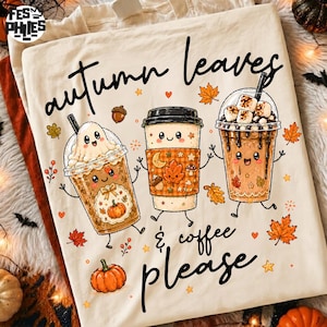 May include: A cream-colored t-shirt featuring a graphic of three anthropomorphic coffee cups with faces. The text reads "autumn leaves & coffee please". The design includes illustrations of a ghost, marshmallows, pumpkins, and orange maple leaves.