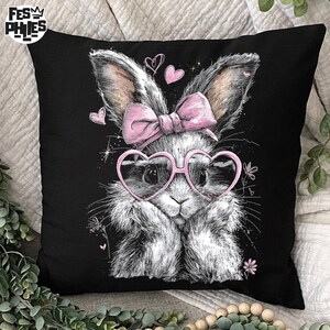 Puede incluir: Coj&iacute;n cuadrado negro con una ilustraci&oacute;n caprichosa de un conejito con gafas en forma de coraz&oacute;n y un lazo rosa. El conejito es blanco y gris con detalles en rosa, rodeado de corazones rosas y detalles florales. El coj&iacute;n tiene el texto "FES PHIES".