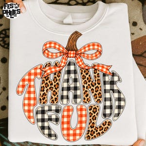 May include: A white crewneck sweatshirt featuring a graphic of a pumpkin shape formed by the word THANKFUL. The letters alternate between red and white buffalo plaid, black and white buffalo plaid, and brown leopard print patterns.