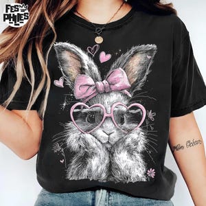 Puede incluir: Camiseta negra con una ilustraci&oacute;n detallada de un conejito con gafas en forma de coraz&oacute;n y un lazo rosa. El conejito tiene un pelaje blanco y gris esponjoso. Corazones rosas y detalles florales rodean el dise&ntilde;o del conejito.