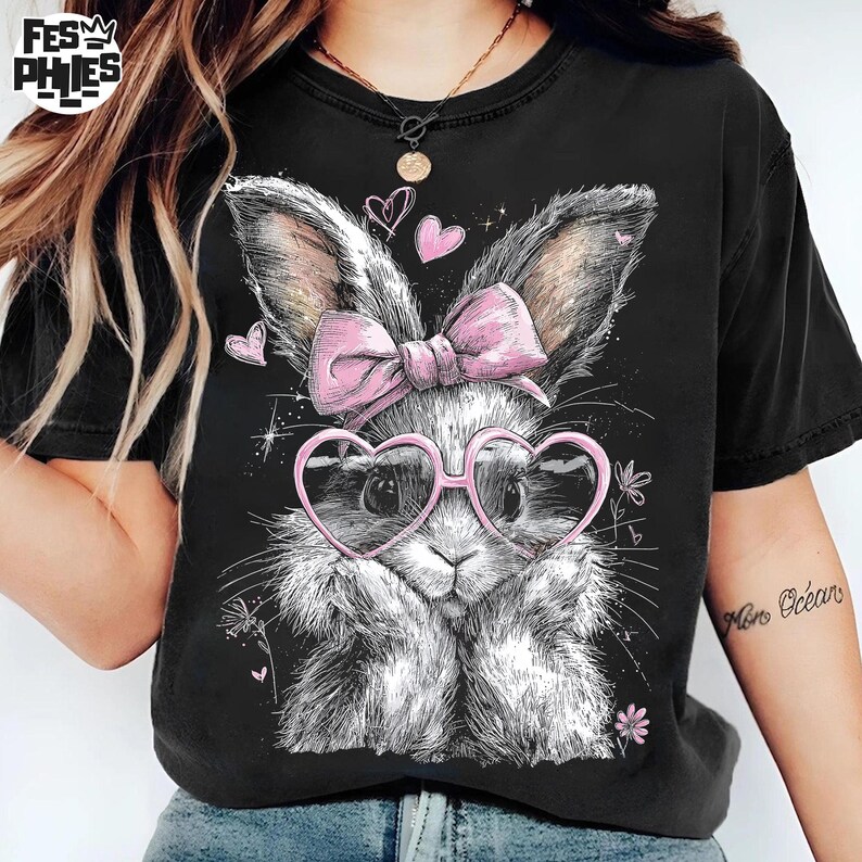 Puede incluir: Camiseta negra con una ilustraci&oacute;n detallada de un conejito con gafas en forma de coraz&oacute;n y un lazo rosa. El conejito tiene un pelaje blanco y gris esponjoso. Corazones rosas y detalles florales rodean el dise&ntilde;o del conejito.
