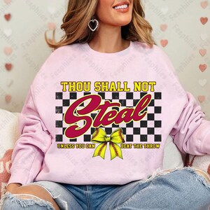 May include: Pink sweatshirt with a black and white checkered background and the text "Thou Shall Not Steal Unless You Can Beat The Throw". A yellow softball with a red bow is in the center of the text.