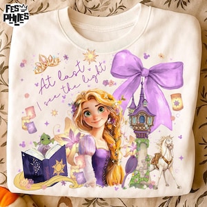 May include: White sweatshirt featuring a colorful illustration of a princess with long blonde hair, a purple dress, and the text "At last I see the light." The design includes a castle, a horse, and a large purple bow.