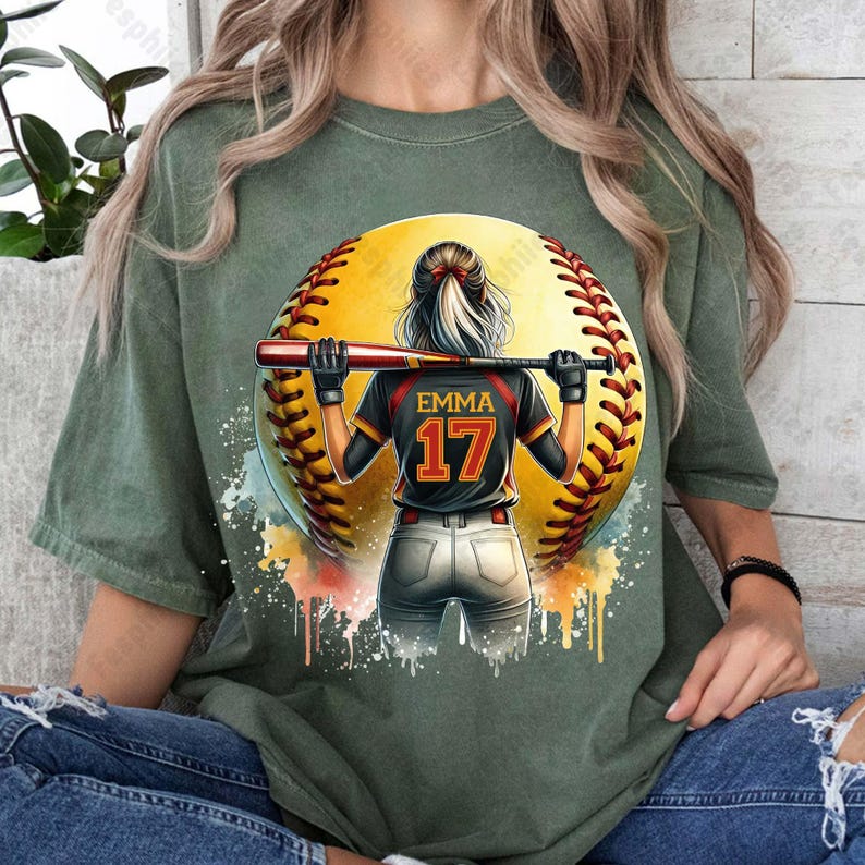 May include: Green t-shirt with a watercolor design of a softball player holding a bat. The player is wearing a black jersey with the name "Emma" and the number "17" on the back. The softball is yellow with red stitching.