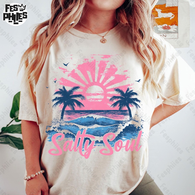 May include: A cream coloured t-shirt with a pink and blue graphic of a sunset over the ocean with palm trees and the text "Salty Soul".