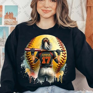May include: A black sweatshirt with a colorful graphic of a softball player holding a bat. The softball is yellow with red stitching and the player is wearing a black jersey with the name "Emma" and the number "17" on the back.