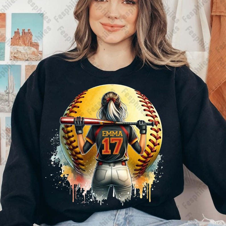 May include: A black sweatshirt with a colorful graphic of a softball player holding a bat. The softball is yellow with red stitching and the player is wearing a black jersey with the name "Emma" and the number "17" on the back.