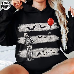 May include: A black hooded sweatshirt featuring a graphic print of a skeleton looking up at a red balloon held by a skeletal hand. The design includes three horizontal distressed white brush strokes, black bat silhouettes, and the text Well, shit...