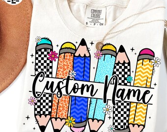 May include: Cream-coloured t-shirt with a colourful pencil design and the words "Custom Name". The pencils feature patterns like checkered, leopard print, and blue zigzags, with floral accents. A gold necklace is draped over the shirt.