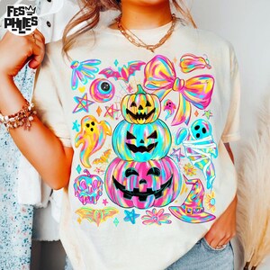 May include: Cream-colored t-shirt featuring a vibrant Halloween-themed design. The graphic includes stacked jack-o'-lanterns, ghosts, bats, and a witch's hat, all in a colorful, tie-dye style. The shirt has a relaxed fit.