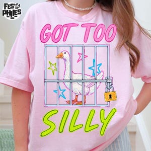 May include: Pink t-shirt with a graphic of a white goose inside a cage, with the words "GOT TOO SILLY" in pink and green. The goose has an orange beak and feet. The cage has a padlock.