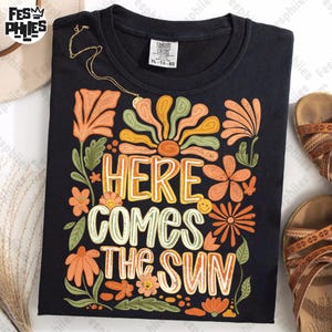 Peut inclure: T-shirt noir avec un motif floral coloré et le texte "Here Comes The Sun".