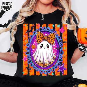 May include: A black t-shirt featuring a graphic of a white ghost wearing a leopard print bow, centered within a purple ornate oval frame. The design includes orange vertical stripes, black bats, colorful maple leaves, and purple stars.