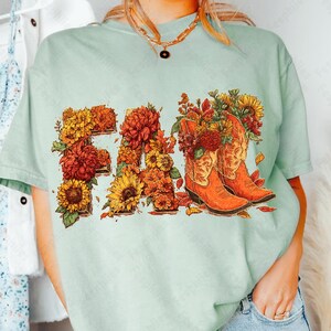 May include: A pale green t-shirt featuring the word "FALL" in a floral design. The letters are filled with sunflowers, chrysanthemums, and autumn leaves. Two orange cowboy boots are filled with flowers. A fall-themed graphic tee.