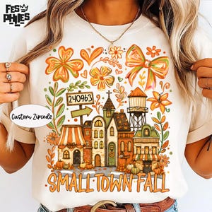 May include: A cream-colored t-shirt featuring a colorful graphic of a small town scene with buildings, a water tower, flowers, and a bow. The text reads Custom Zipcode, 240963, and SMALL TOWN FALL. The design includes orange, green, and brown tones.