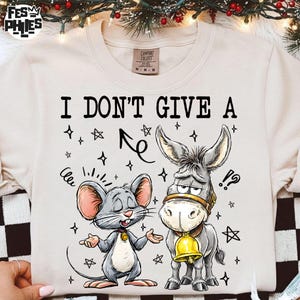 May include: Cream-colored sweatshirt with the phrase "I DON'T GIVE A" above a cartoon mouse and donkey. The mouse has its hands out, and the donkey wears a bell. The design includes stars and an arrow.