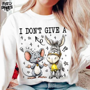 May include: White sweatshirt with the text "I DON'T GIVE A" above a cartoon illustration of a mouse and a donkey. The mouse is gray with pink ears, and the donkey is gray with a gold bell.
