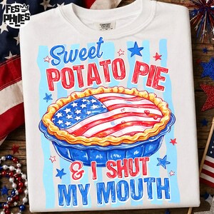 May include: White t-shirt with a graphic of a sweet potato pie decorated with an American flag design. The text reads "Sweet Potato Pie & I Shut My Mouth" in red and blue. The shirt has a crew neck.