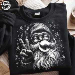 May include: Black sweatshirt with a white graphic of Santa Claus wearing sunglasses and blowing a bubble. Santa is giving the peace sign, surrounded by snowflakes. The sweatshirt has the text "FESM PHIES" on the upper left.