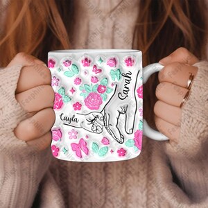 May include: White ceramic mug with a pink and green floral design. The mug features two hands, one with the name "Sarah" and the other with the name "Cayla".