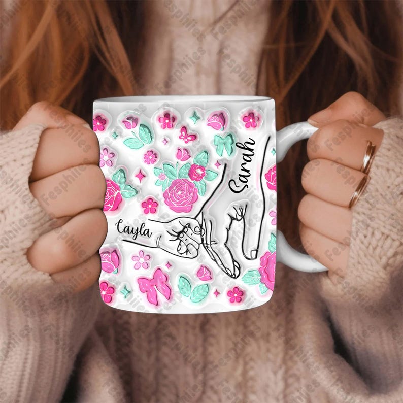 May include: White ceramic mug with a pink and green floral design. The mug features two hands, one with the name "Sarah" and the other with the name "Cayla".
