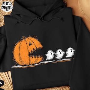 May include: Black hoodie featuring a Halloween-themed graphic. An orange jack-o'-lantern with a jagged mouth is chasing three white ghosts. The design is in a cartoon style, perfect for the spooky season.