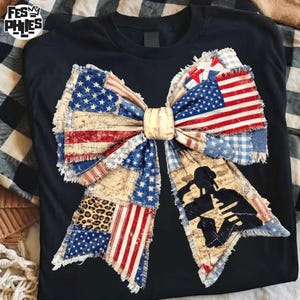 Könnte beinhalten: Schwarzes T-Shirt mit einem großen Schleifendesign aus Stoffflicken im amerikanischen Flaggen-Design, Leopardenmuster und Vintage-Schrift. Die Schleife hat einen goldfarbenen Mittelknoten und eine Silhouette von zwei Soldaten.