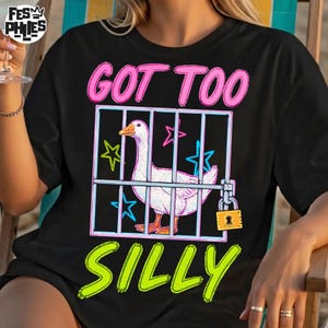 May include: Black t-shirt with a graphic of a white goose inside a cage, the text "GOT TOO SILLY" in pink and green. The cage has a padlock and star accents.