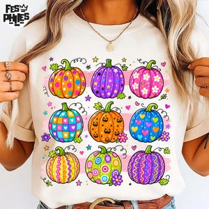 Op de afbeelding: Een cr&egrave;mekleurig t-shirt met een grafische print van negen pompoenen in een raster. Elke pompoen heeft een uniek patroon, waaronder stippen, sterren, bloemen, hartjes, pompoengezichten en krullen in levendige kleuren.