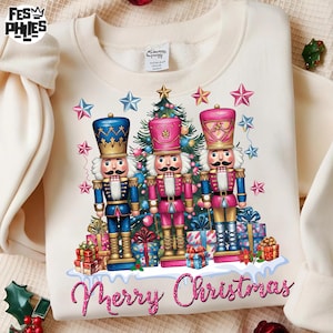 May include: Cream-colored sweatshirt featuring a festive design with three nutcracker figures in blue, pink, and gold uniforms. A Christmas tree and star accents surround the nutcrackers, with the words "Merry Christmas" in pink script at the bottom.