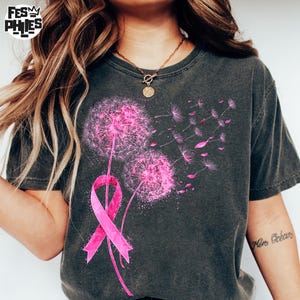 May include: Dark gray t-shirt featuring a pink dandelion design with a pink ribbon. The dandelion has fluffy seed heads and seeds blowing away. The shirt has a crew neck and short sleeves.