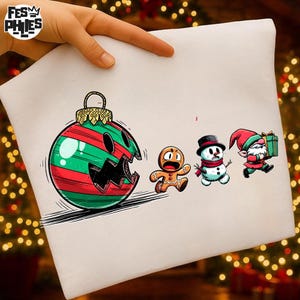 Könnte beinhalten: Ein weißes Kissenbezug mit einem Cartoon-Weihnachtsdesign. Eine große grün-rot gestreifte Christbaumkugel mit offenem Mund jagt einen Lebkuchenmann, einen Schneemann und einen Elf. Der Kissenbezug hat den Text "FES PHIES" in der oberen linken Ecke.