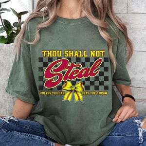 May include: Green t-shirt with a black and white checkered background and the text "Thou Shall Not Steal" in red. A yellow softball with a red bow is in the center of the design. The text "Unless You Can Beat The Throw" is below the softball.