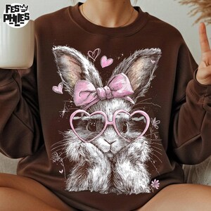 Puede incluir: Sudadera marr&oacute;n con un dise&ntilde;o de un conejo blanco y gris con gafas de coraz&oacute;n y un lazo rosa. El dise&ntilde;o incluye corazones y flores. La sudadera tiene cuello redondo y mangas largas, ideal para uso diario.