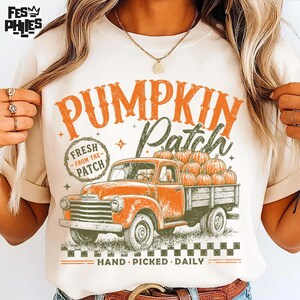 May include: A cream-colored t-shirt featuring a graphic of an orange vintage pickup truck loaded with pumpkins. The text reads PUMPKIN Patch, FRESH FROM THE PATCH, and HAND PICKED DAILY. The design includes orange and green accents with a checkered border.