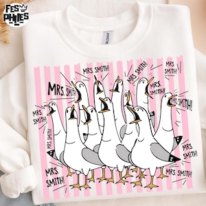 May include: White sweatshirt featuring a cartoon illustration of seagulls with open beaks, set against a pink striped background. The text "MRS. SMITH!" is repeated around the seagulls. The sweatshirt has a crew neck and long sleeves.