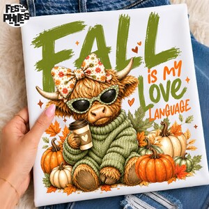 May include: A white t-shirt featuring a graphic of a fluffy highland cow wearing a floral bow and black sunglasses, holding a coffee cup. The text reads FALL is MY Love LANGUAGE. The design includes orange pumpkins and green autumn leaves.