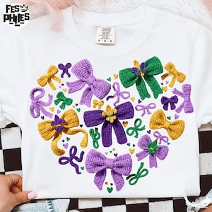 May include: White t-shirt featuring a heart-shaped design of crocheted bows in purple, green, and gold. The shirt has the text "Fes Philes" at the top left and a "Comfort Colors" tag.
