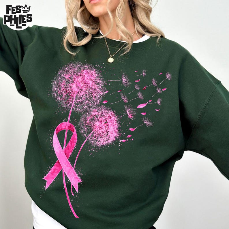 May include: Dark green sweatshirt featuring a pink dandelion design with a pink ribbon. The dandelion is in full bloom with seeds blowing away. The sweatshirt has a crew neck and long sleeves. The design is centered on the front of the shirt.